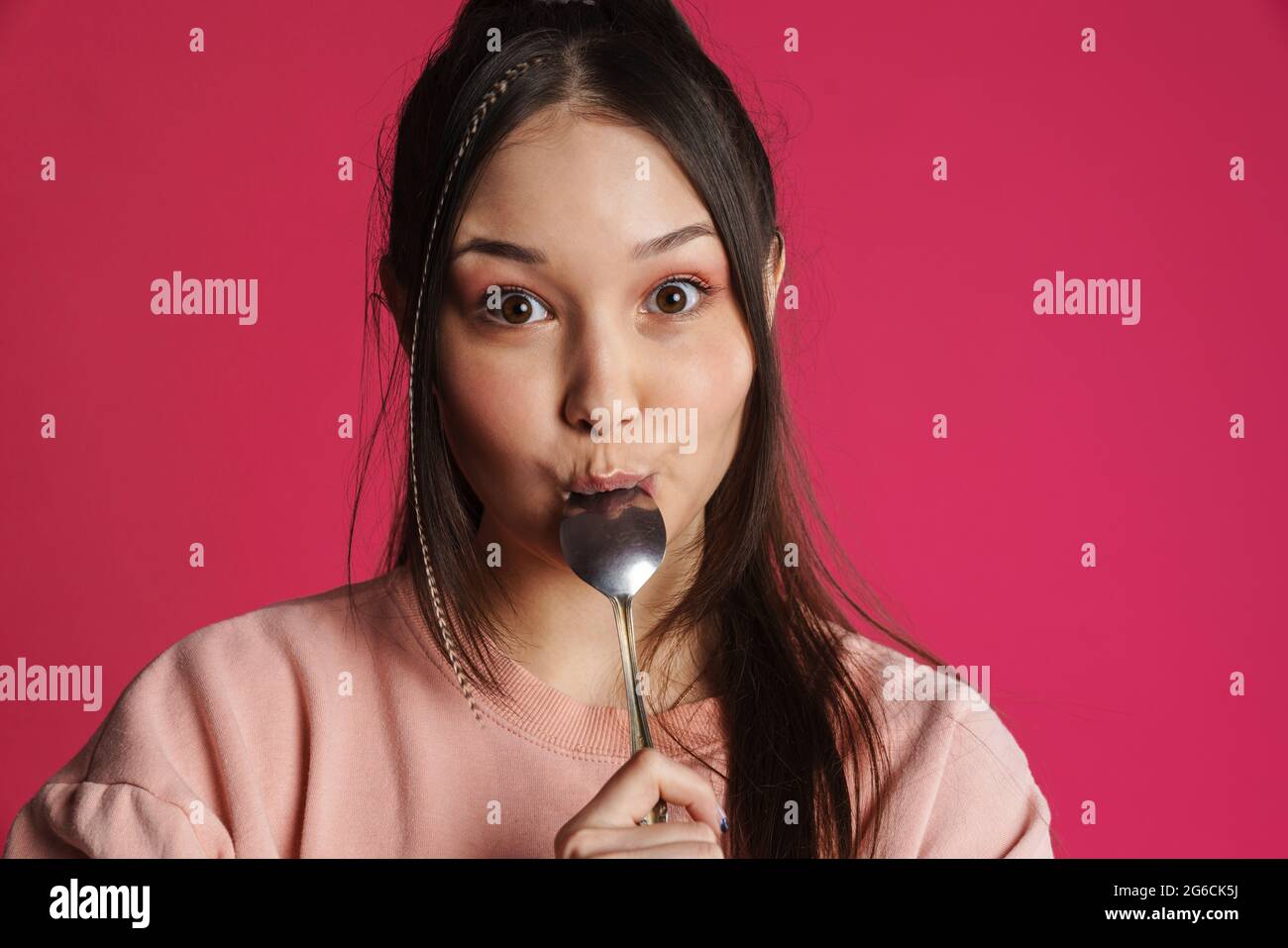 Asian brunette donna felice leccando il suo cucchiaio e guardando la macchina fotografica isolata sopra la parete viola Foto Stock