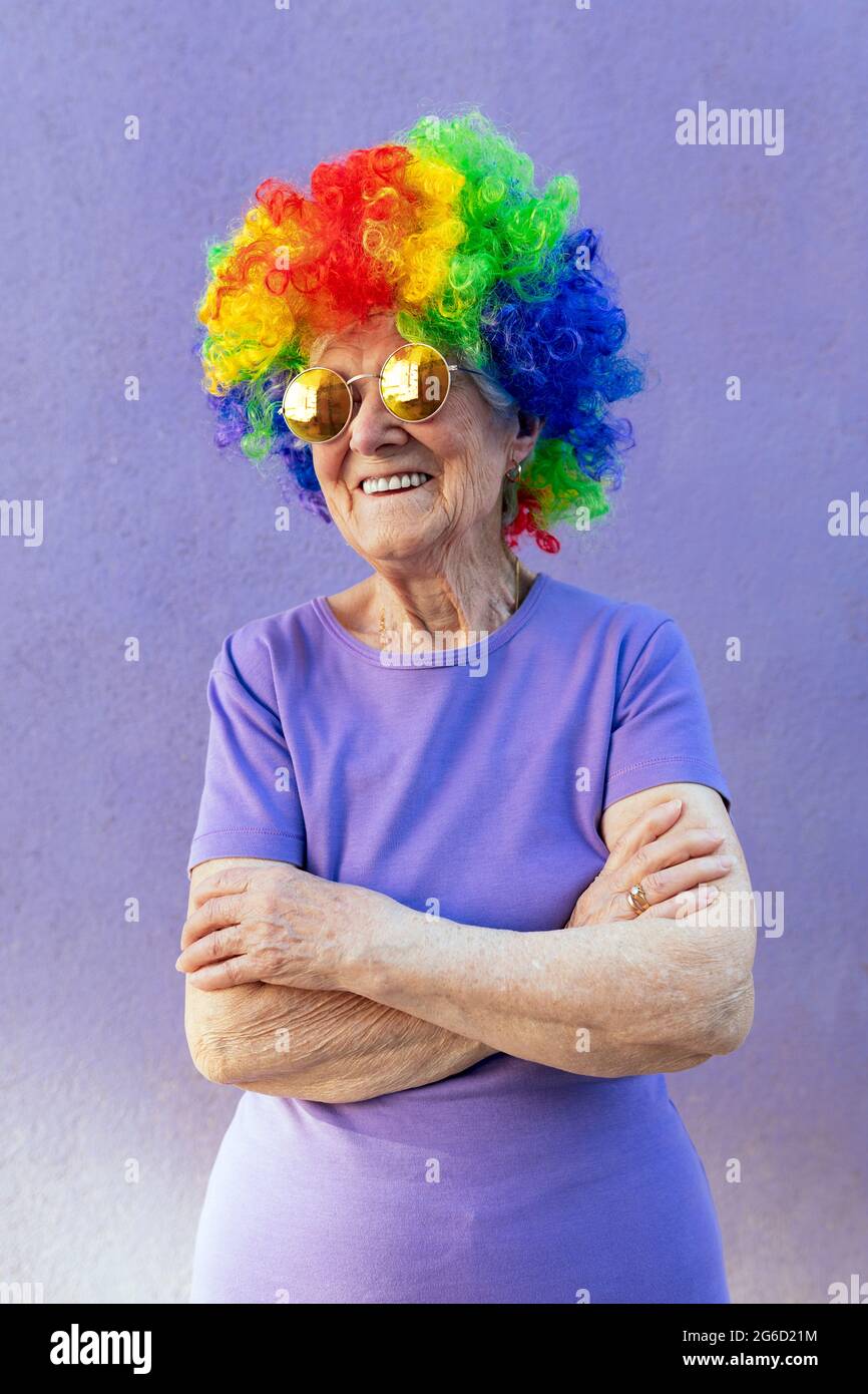 Contenuto atleta senior femminile in moderni occhiali da sole e parrucca brillante in piedi con braccia ripiegate su sfondo viola Foto Stock
