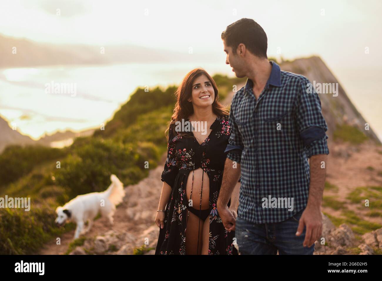Happy Hispanic incinta Donna e l'uomo amorevole tenendo le mani e guardandosi mentre camminando con il cane lungo la costa collinare in estate sera Foto Stock