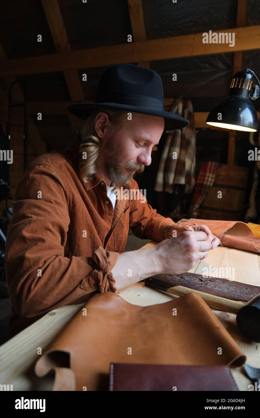 Giovane lavoratore in cappello seduto sul suo posto di lavoro e facendo scarpe da cuoio in officina Foto Stock