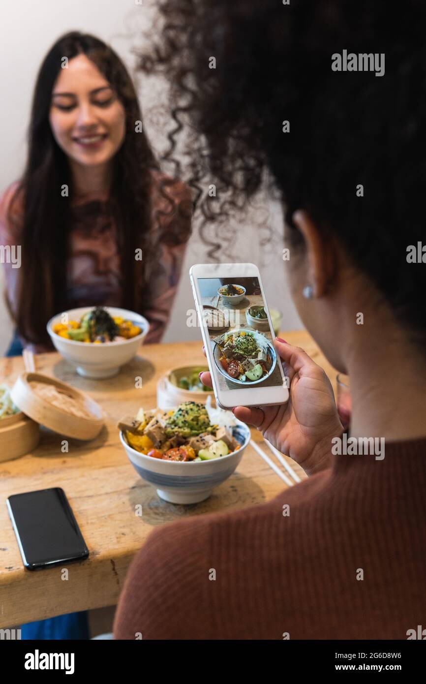 Dall'alto di Unriconoscable Woman scattare foto di piatto di poke su smartphone mentre si passa il fine settimana in ristorante con un amico femmina Foto Stock