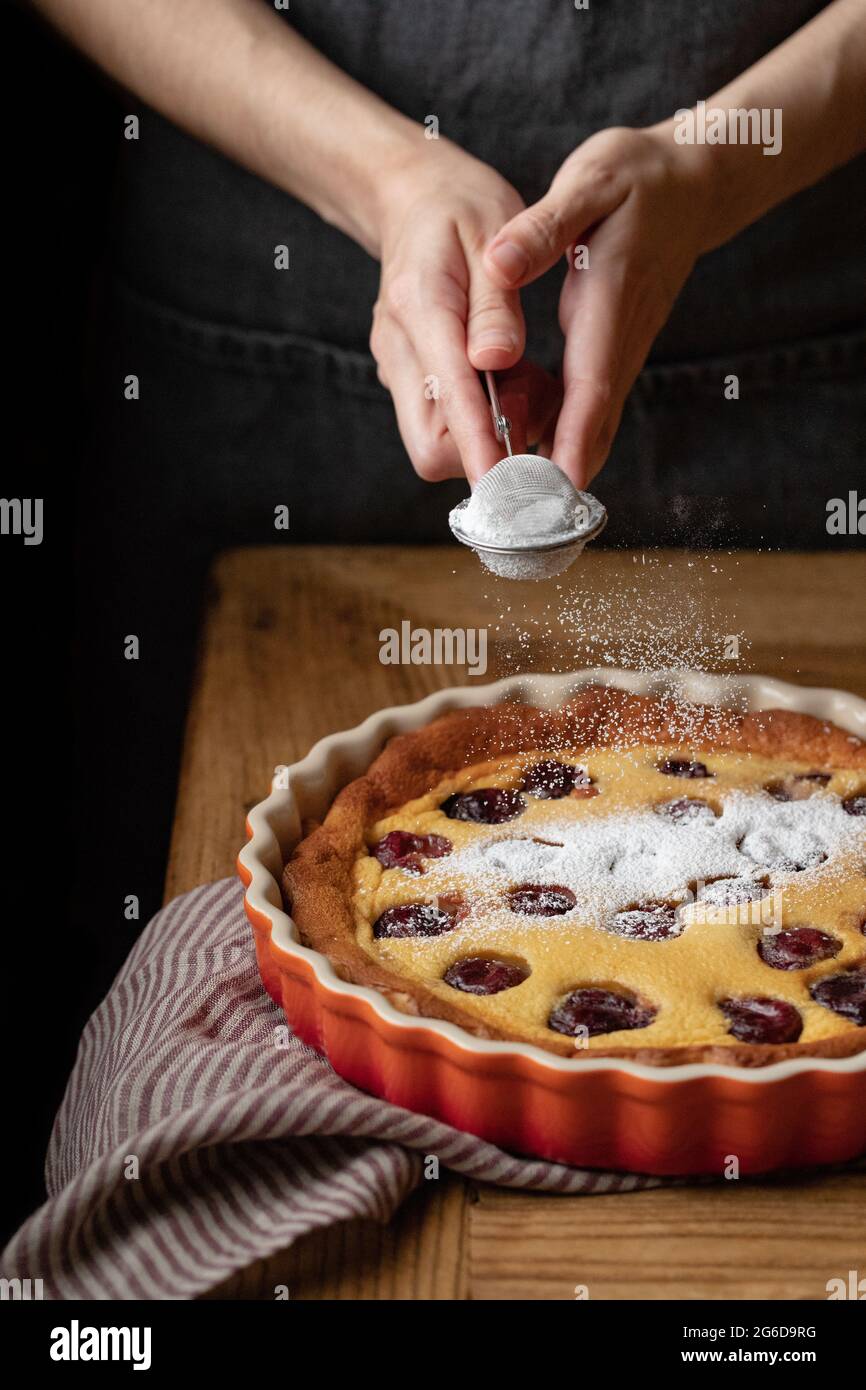 Persona anonima in grembiule in piedi a tavola di legno e polvere di zucchero spruzzante in cima alla deliziosa torta fatta in casa con ciliegie Foto Stock