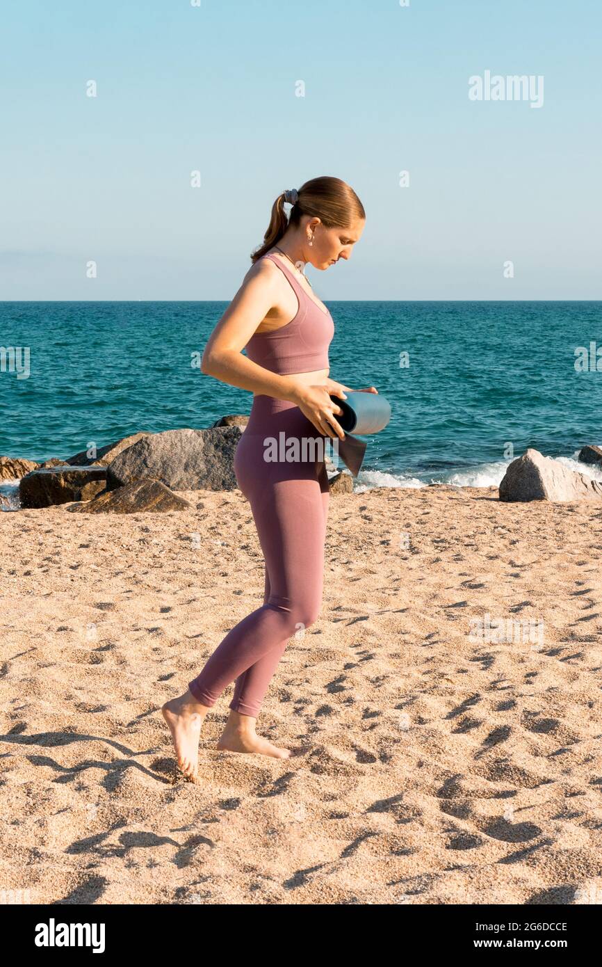 Vista laterale del corpo della giovane femmina in abbigliamento sportivo che mette un tappetino yoga sulla sabbia mentre si prepara per la pratica sulla spiaggia vicino all'oceano Foto Stock