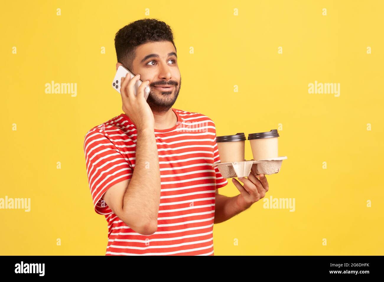 Uomo indaffarato in t-shirt rossa a righe che porta il telefono nel porta-mano con due tazze da caffè, concetto di consegna rapida e facile da ordinare. Riprese in studio al coperto Foto Stock