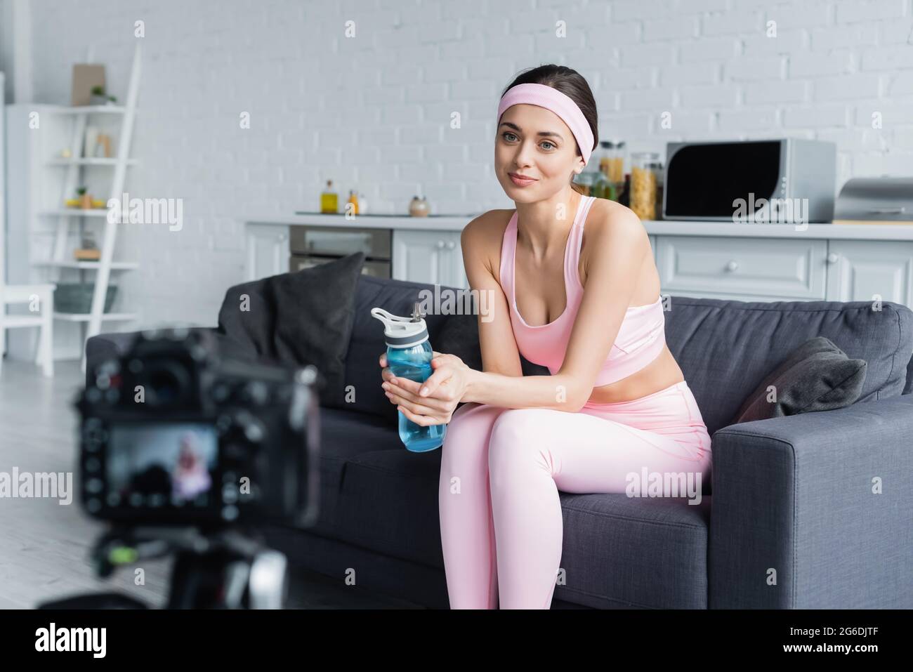 donna sorridente seduta con una bottiglia sportiva davanti a una fotocamera digitale sfocata Foto Stock