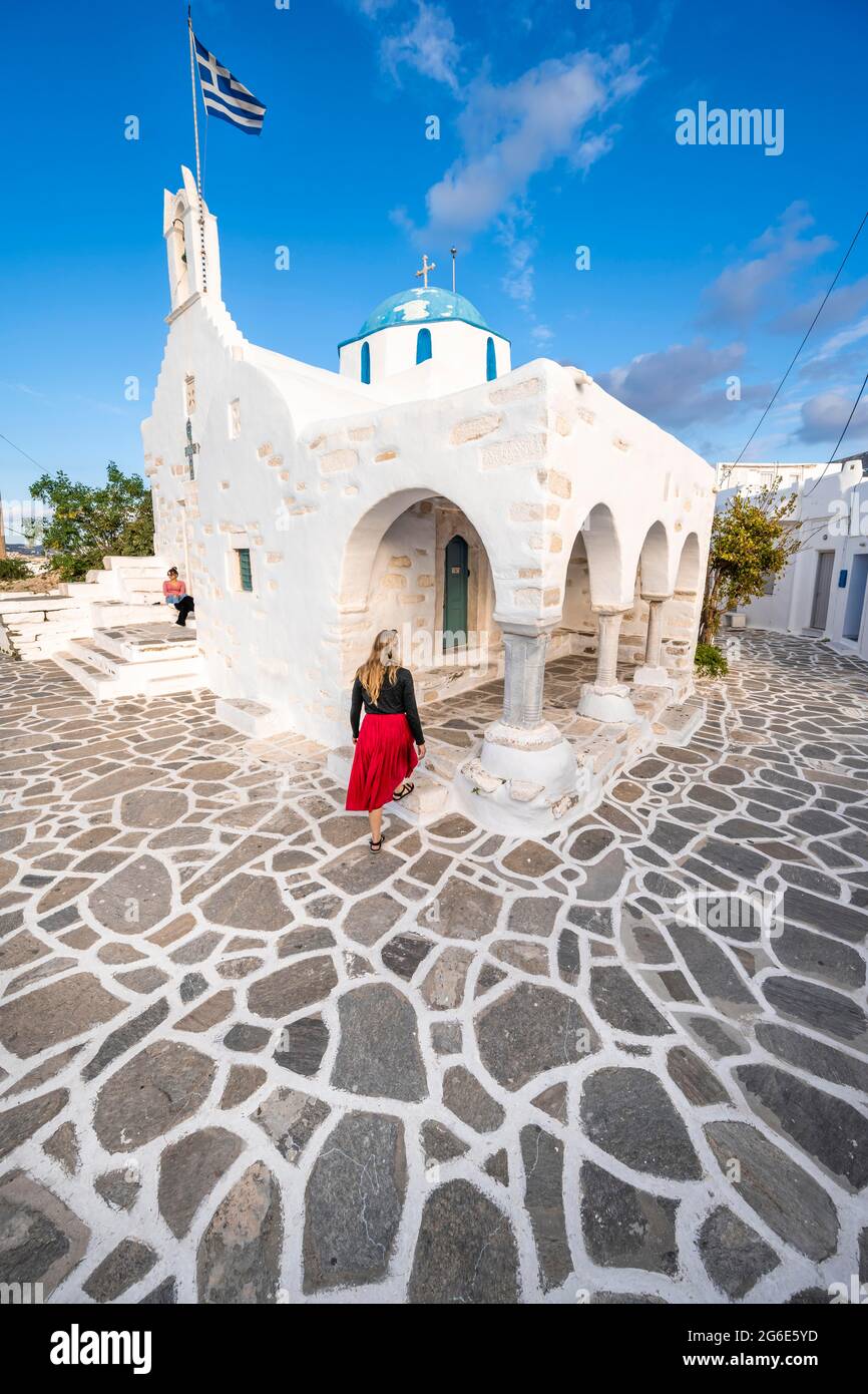 Turismo con abito rosso, blu e bianco greco ortodosso Chiesa Agios Nikolaos con bandiera greca, Parikia, Paros, Cicladi, Mar Egeo, Grecia Foto Stock