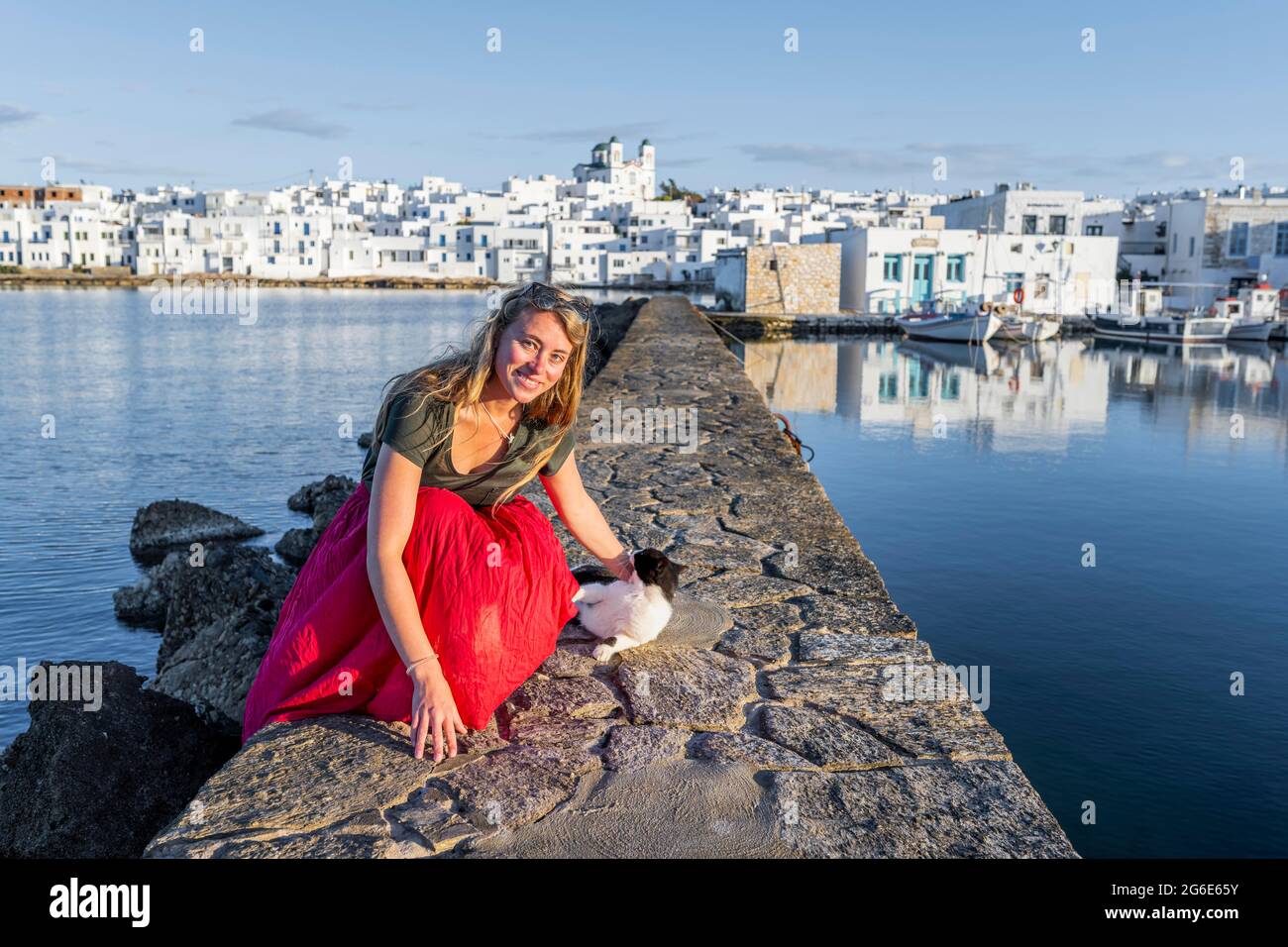 Gatto e giovane donna con abito rosso sulla parete del porto, dietro la chiesa di Naoussa, città portuale Naoussa, isola Paros, Cicladi, Grecia Foto Stock