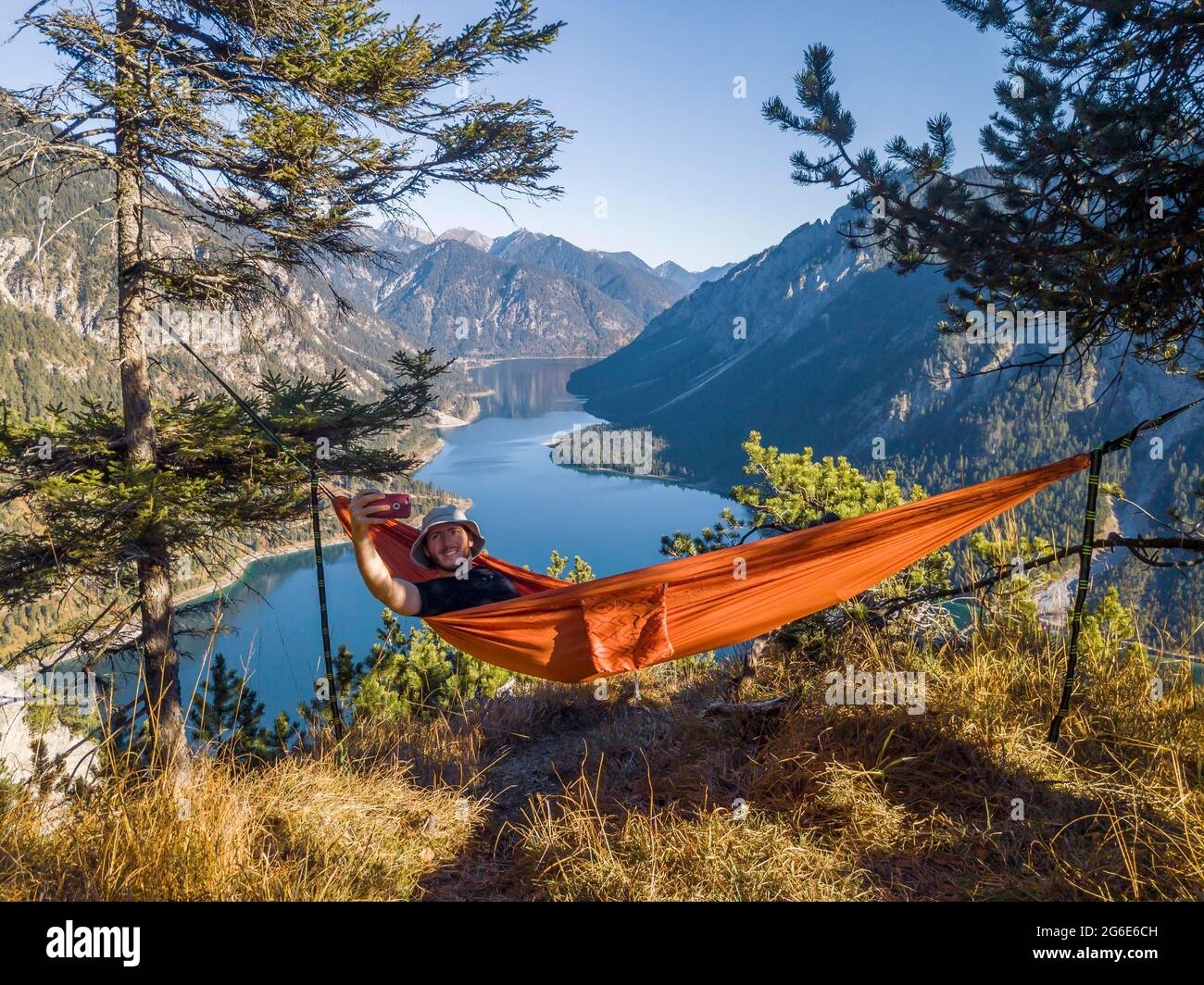 Uomo che scatta foto di se stesso con il telefono cellulare, escursionista seduto in amaca con vista sulle montagne con lago, Plansee, Alpi Ammergau, quartiere Reutte Foto Stock