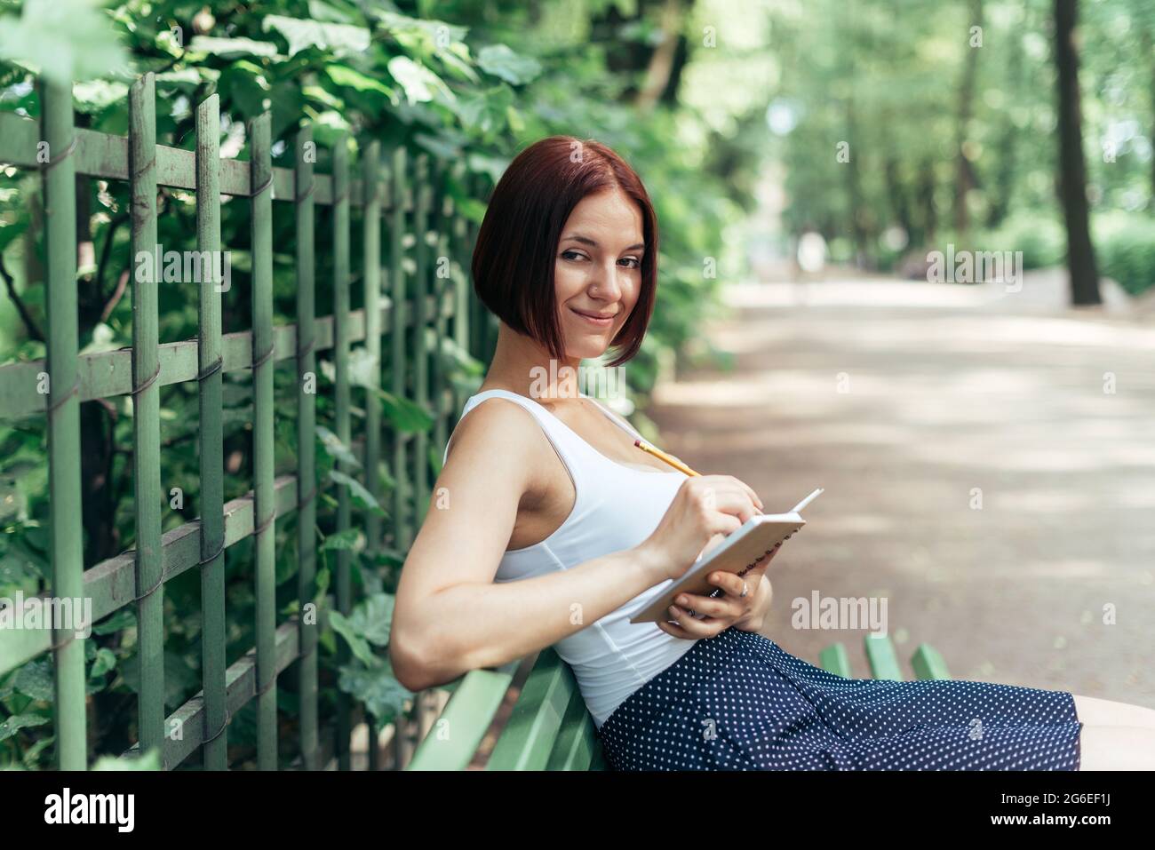 La ragazza caucasica dai capelli rossi si siede su una panchina in un parco estivo e prende appunti in un notebook. Foto Stock