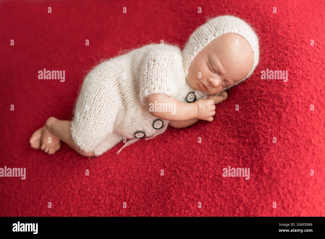 Bambola neonato che indossa un jumpsuit in maglia bianca e un cappello sul  cofano in maglia che giace su un lato su una coperta rossa. Foto per  bambini primo natale, nessun modello