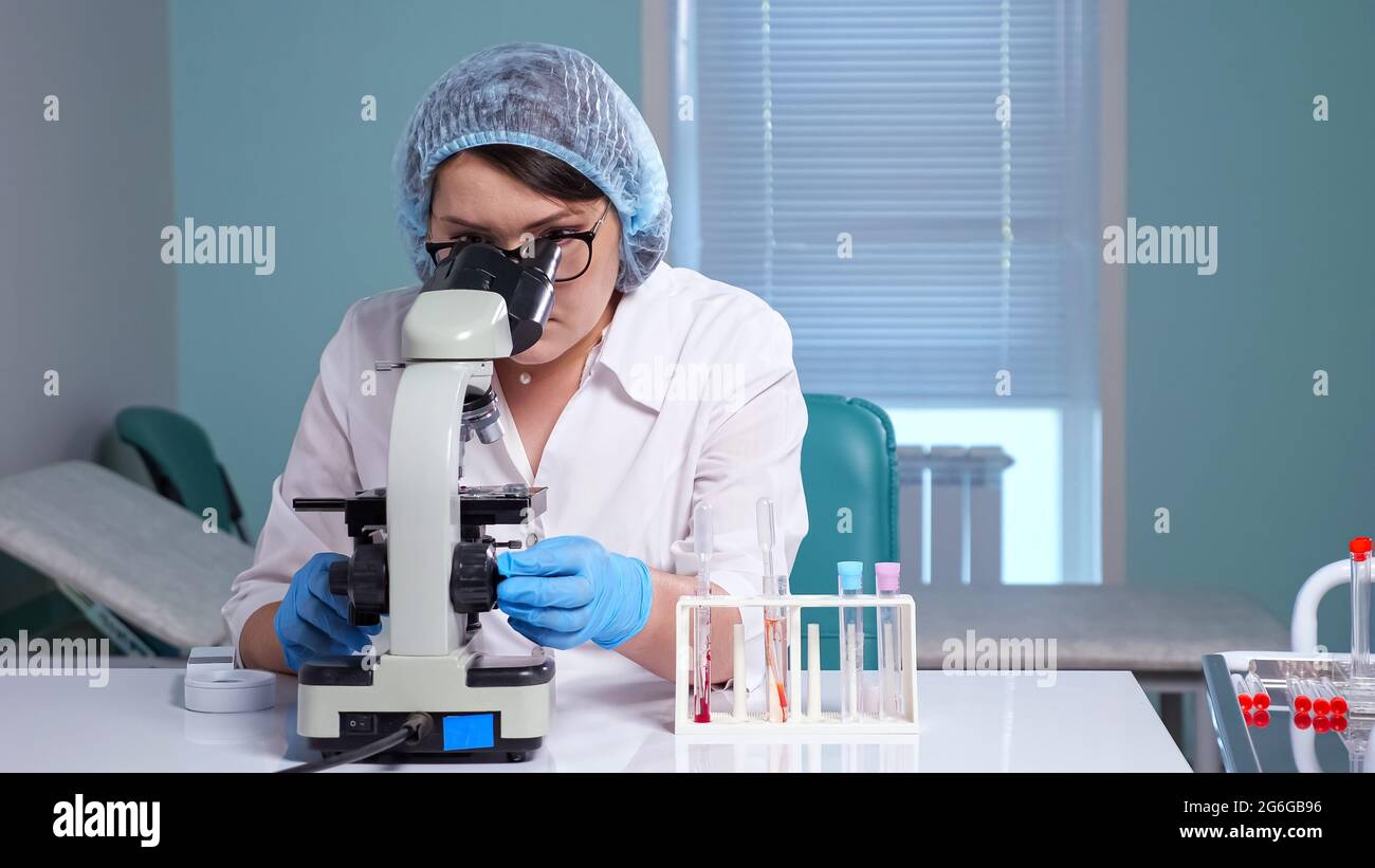 Giovane donna in occhiali e uniforme bianca guarda il campione attraverso un microscopio contemporaneo seduto sul posto di lavoro in laboratorio clinico Foto Stock