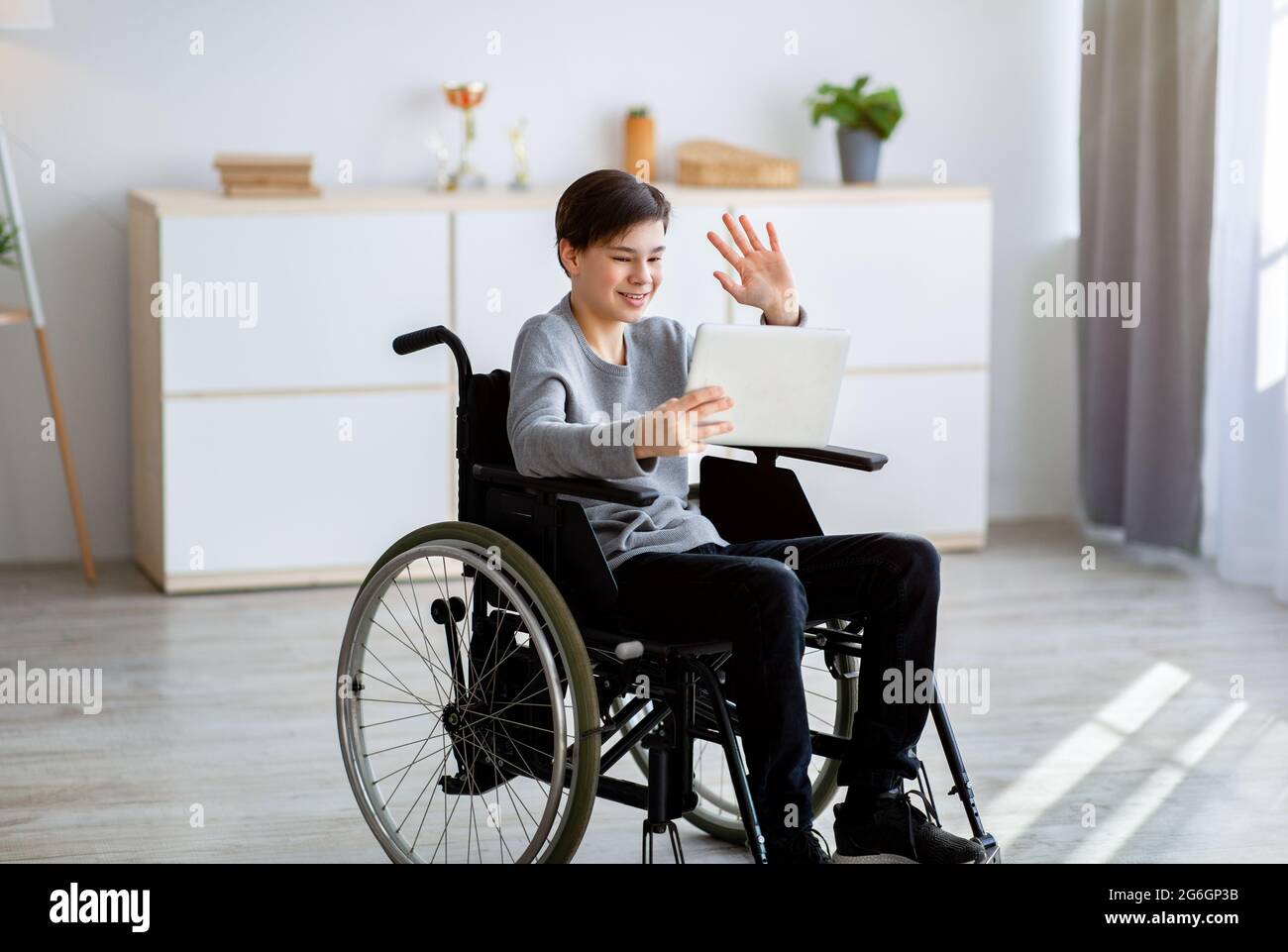 Un adolescente in sedia a rotelle con handicap positivo che ha una videoconferenza con l'insegnante su un tablet pc, agitando sullo schermo all'interno Foto Stock