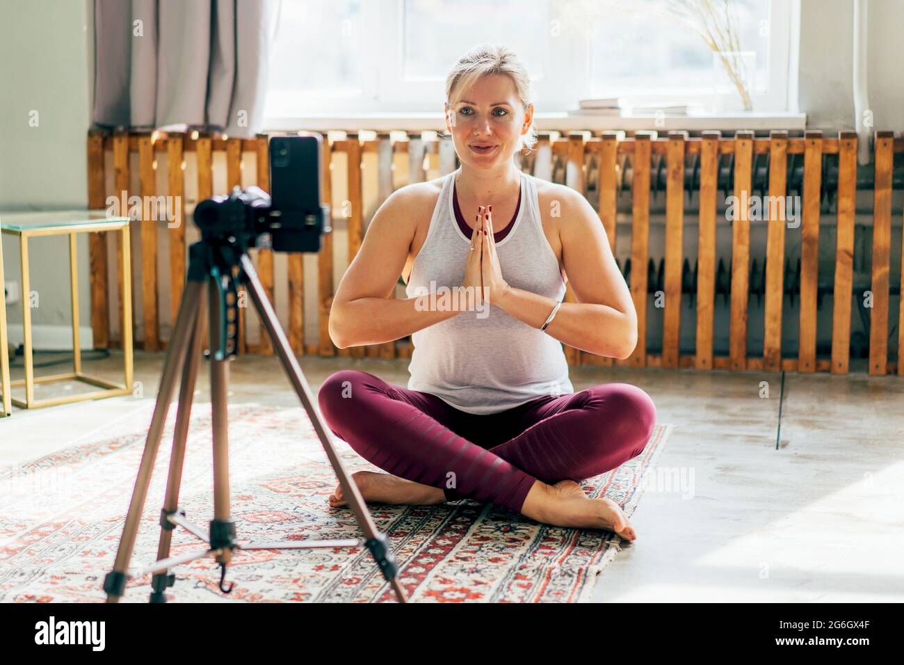 Una donna seduta sul pavimento in una posizione lotus e salutata con un gesto namaste guarda lo schermo del telefono. Comunicazione remota nei social media e. Foto Stock