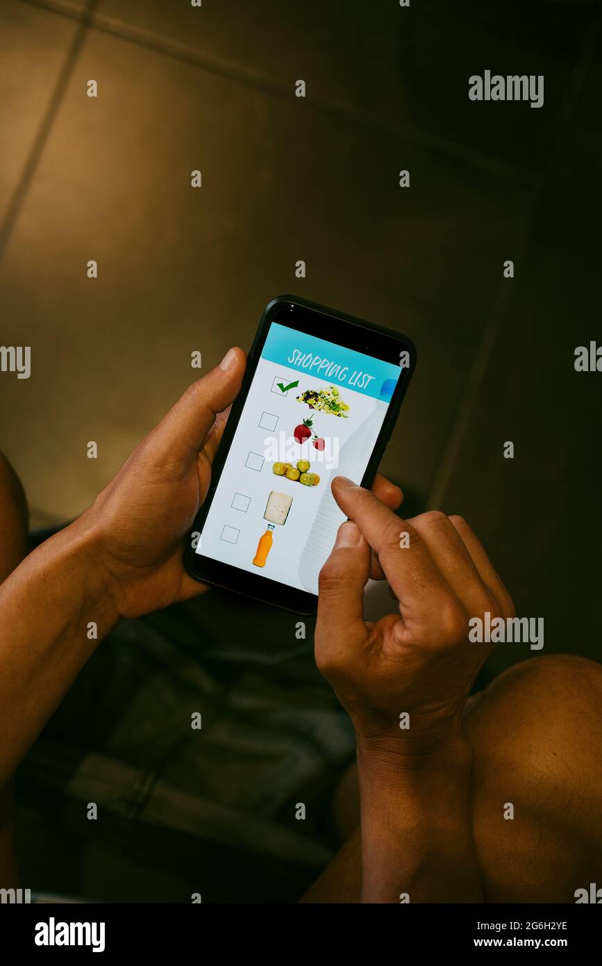 un giovane caucasico, seduto in toilette, sta facendo la sua lista di negozi nel suo smartphone, con un'app simulata con immagini di prodotti diversi Foto Stock