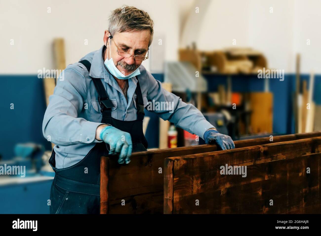 Un uomo anziano dipinge tavole di legno con vernice scura in un laboratorio di Falegnameria. Un pensionato in una maschera medica guadagna un lavoro part-time come un cabinetmaker. Lavoro fisico Foto Stock