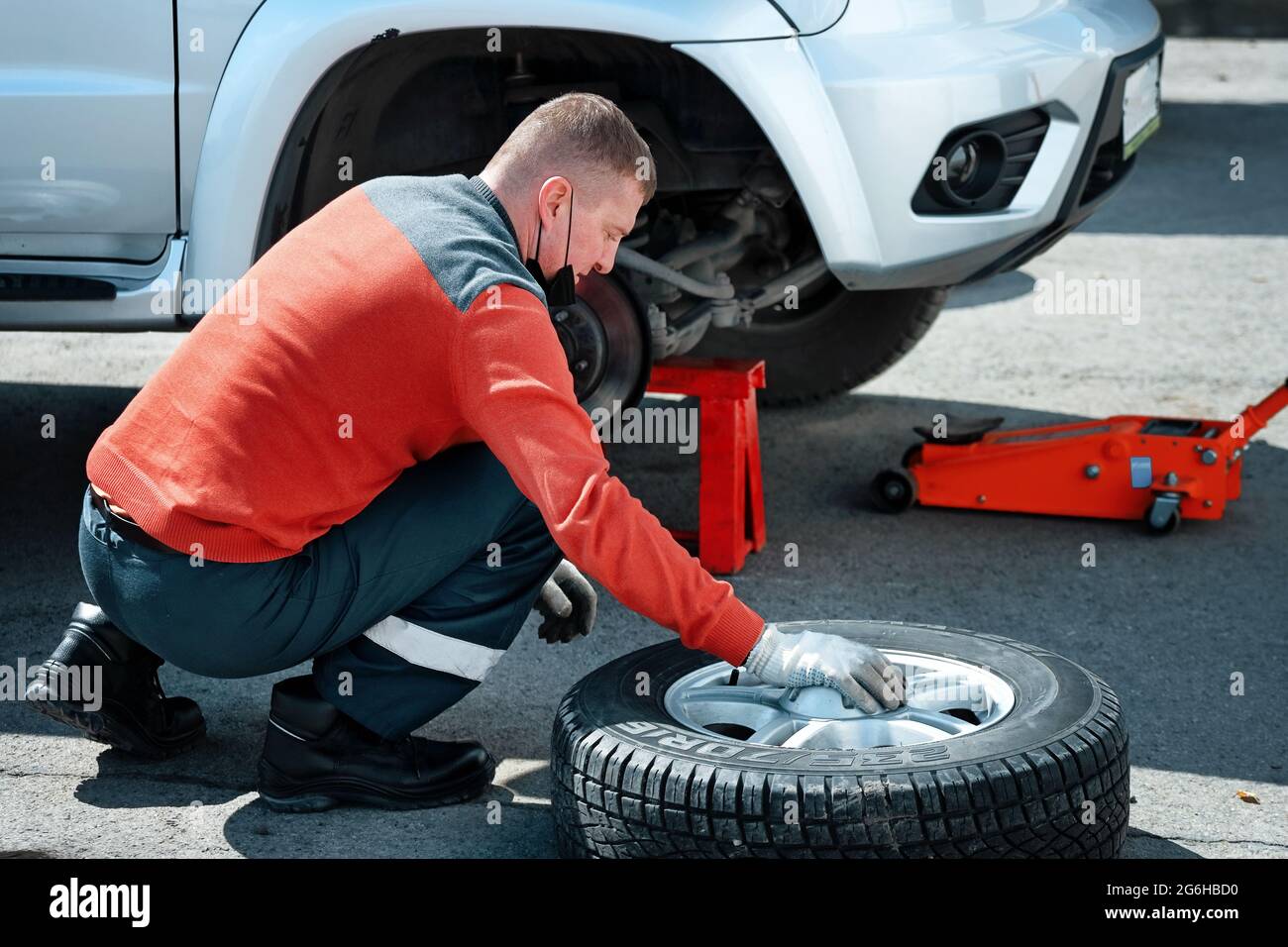 Un meccanico di automobile in una maschera medica cambia una ruota su un automobile sulla strada in una giornata soleggiata. Guida su strada con una foratura degli pneumatici. Manutenzione urgente degli pneumatici Foto Stock