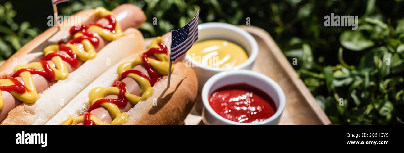 hot dog con piccola bandiera degli stati uniti vicino a ciotole con ketchup e senape su prato verde, banner Foto Stock