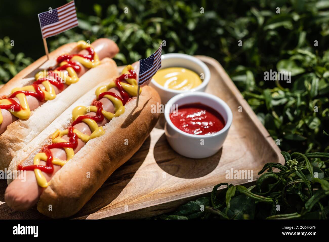 hot dog con piccole bandiere usa vicino ketchup e senape su vassoio di legno e erba verde Foto Stock