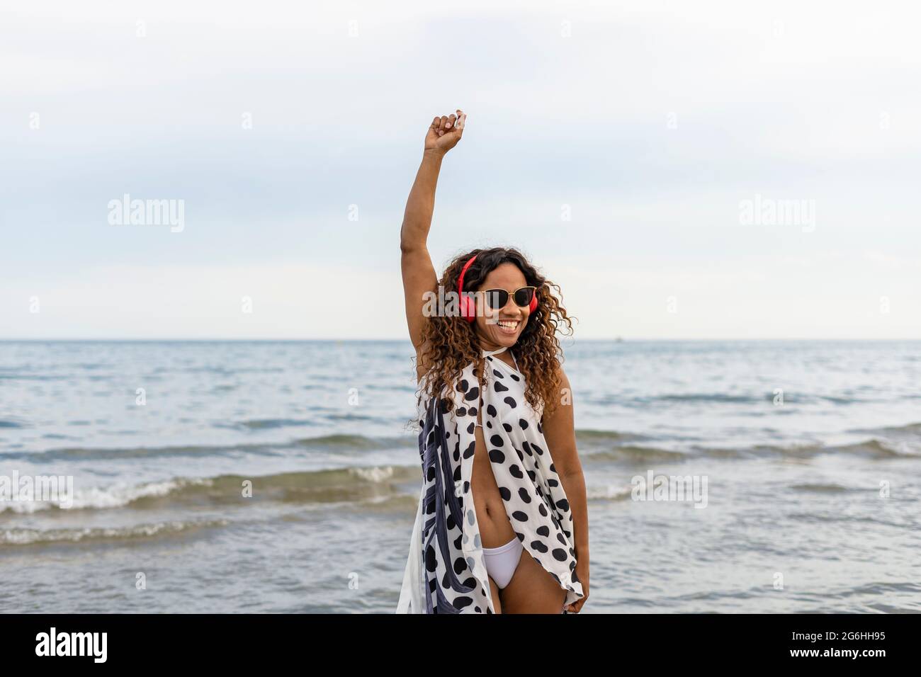 Felice giovane bella donna afro americana sorridente, indossando cuffie danzanti e alzando il braccio sulla spiaggia.Femminile godendo di vacanze estive concetto li Foto Stock