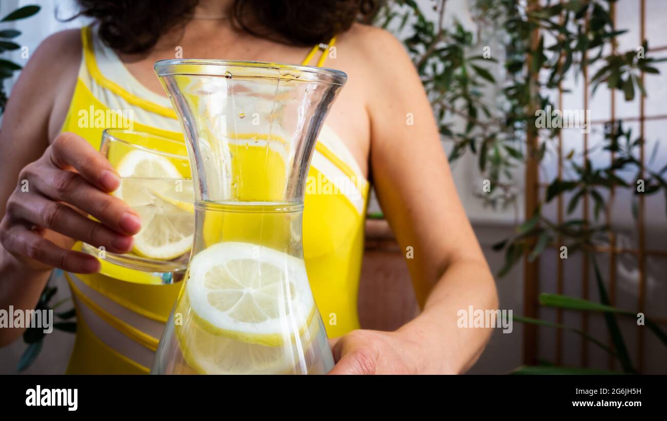 Donna in un vestito estivo giallo con una bottiglia d'acqua offre un bicchiere d'acqua con una fetta di limone succosa. Primo piano. Foto Stock