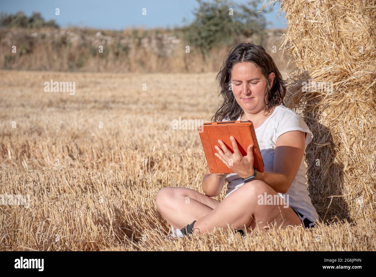 Donna latina bruna di mezza età seduta a terra in un campo appoggiato su un mucchio di balle di paglia utilizzando una tavoletta con copertura arancione. Tecnologia An Foto Stock