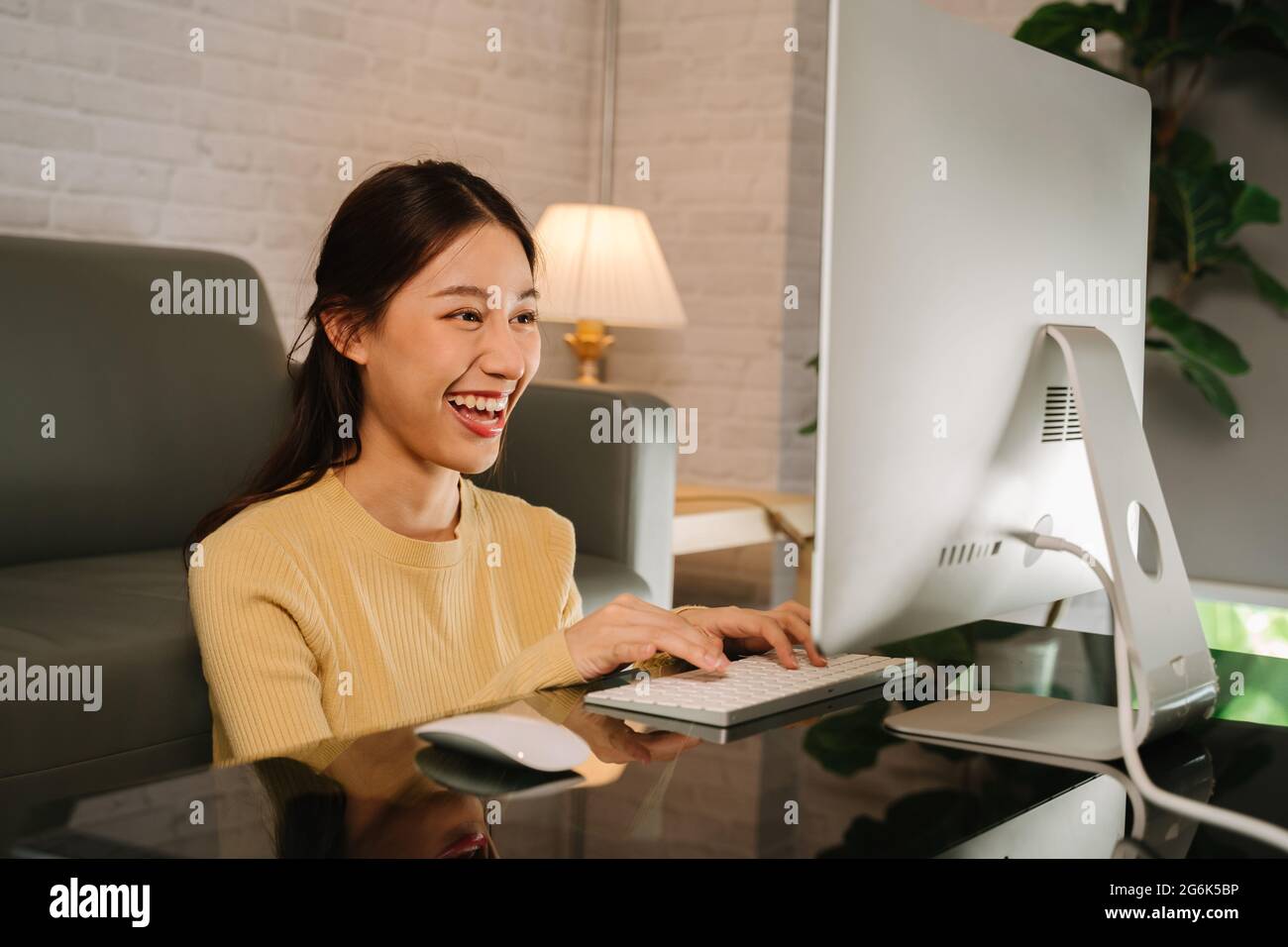 Felice giovane donna d'affari asiatica di successo che lavora da casa e che scrive sulla tastiera usando il computer desktop mentre si siede in soggiorno Foto Stock