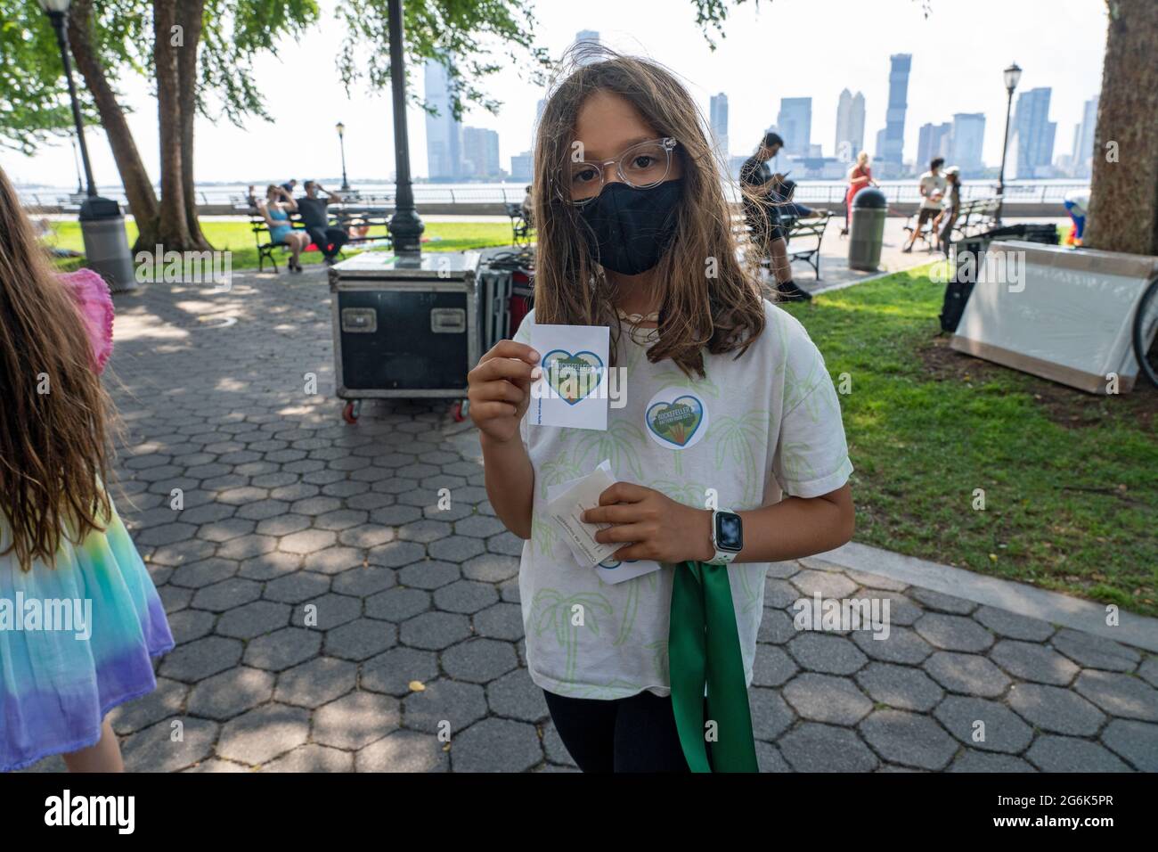 Un bambino consegna adesivi e nastri verdi durante una protesta contro i piani del governatore Andrew Cuomo per un memoriale COVID-19 a New York City. Duecento manifestanti si sono radunati nel Rockefeller Park di Lower Manhattan, cercando di mantenere la pressione sul governatore Andrew Cuomo pochi giorni dopo i potenti e. il leader di stato scandaloso ha nixed i suoi piani di spianare una porzione dello spazio verde per il monumento, il memoriale del coronavirus, il 'cerchio degli eroi'. La protesta di Battery Park City si è verificata come il Consiglio della Comunità 1, che rappresenta l'area, ha programmato una riunione di mercoledì per discutere il monumento. ( Foto Stock