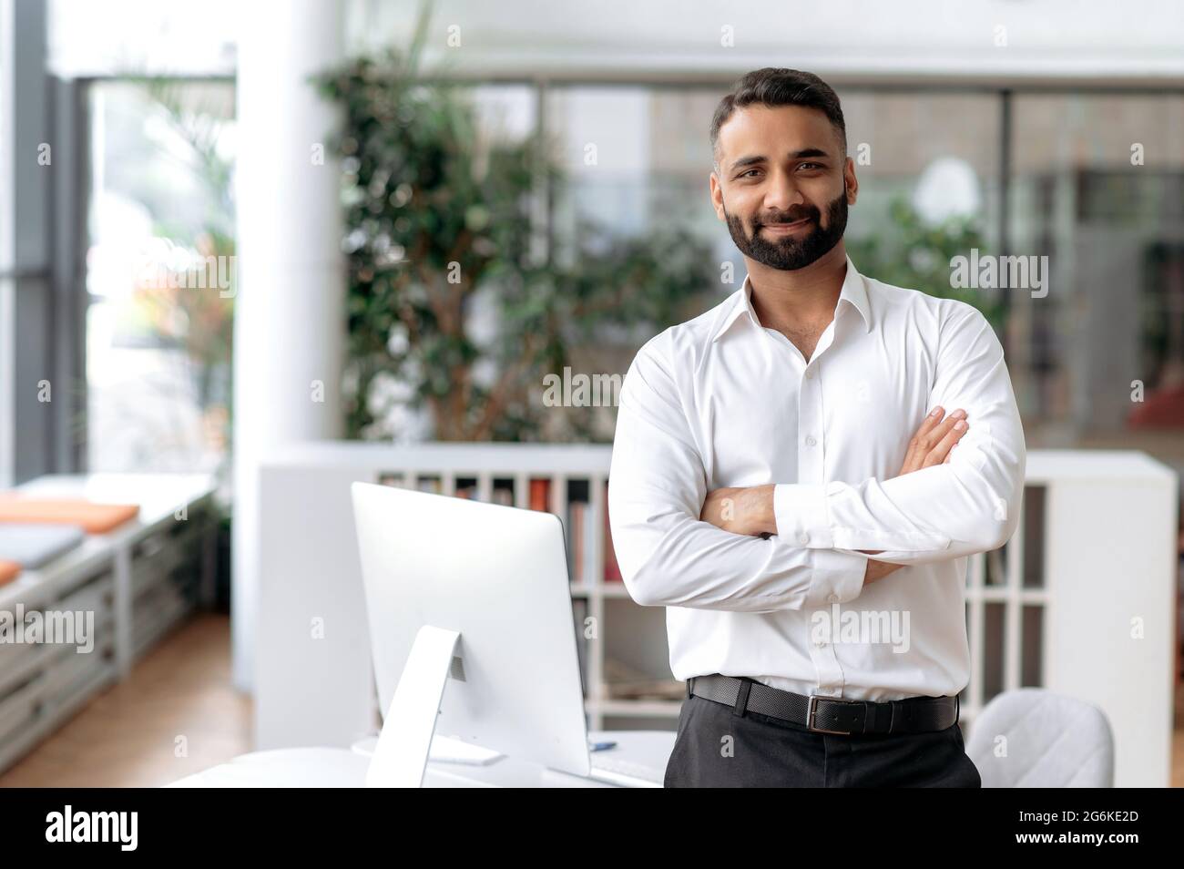 Ritratto formale di uomo d'affari. Un uomo d'affari indiano sicuro di successo o manager, in camicia bianca, si trova vicino alla sua scrivania in ufficio, le braccia incrociate, guarda direttamente la fotocamera e sorride amichevole Foto Stock