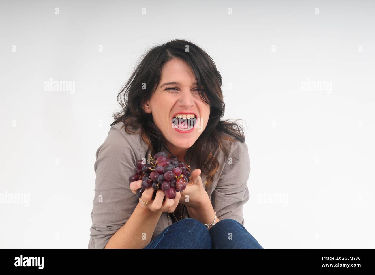 esagerato e umoristico sguardo a una giovane donna che mangia uva da uno studio di uva rossa girato su sfondo bianco Foto Stock