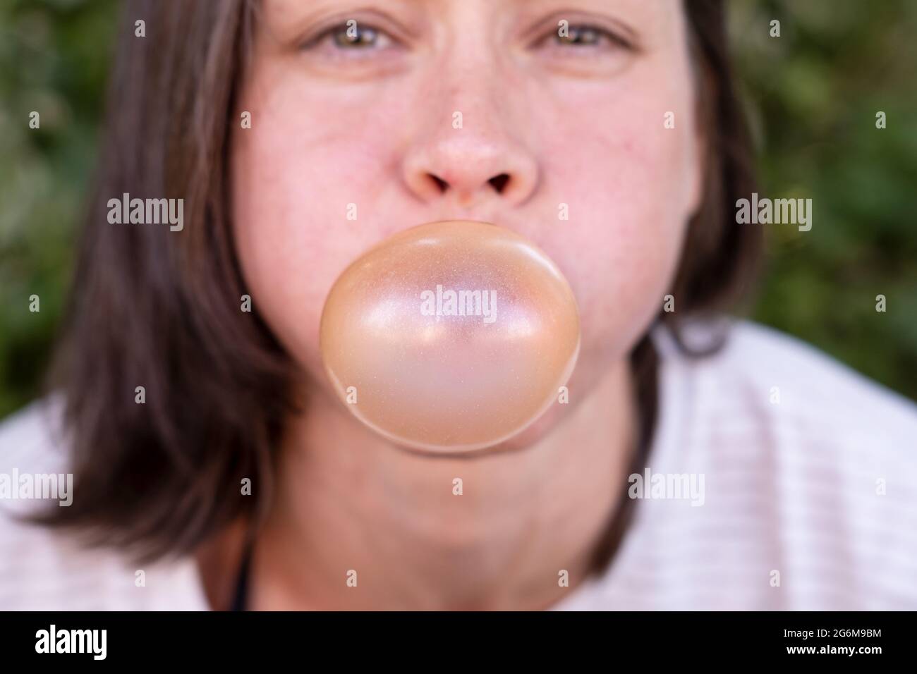 Closeup ritratto di donne divertenti, gonfia con cura la gomma bolla, con gli occhi stretti e le guance rotonde, all'aperto. Foto Stock