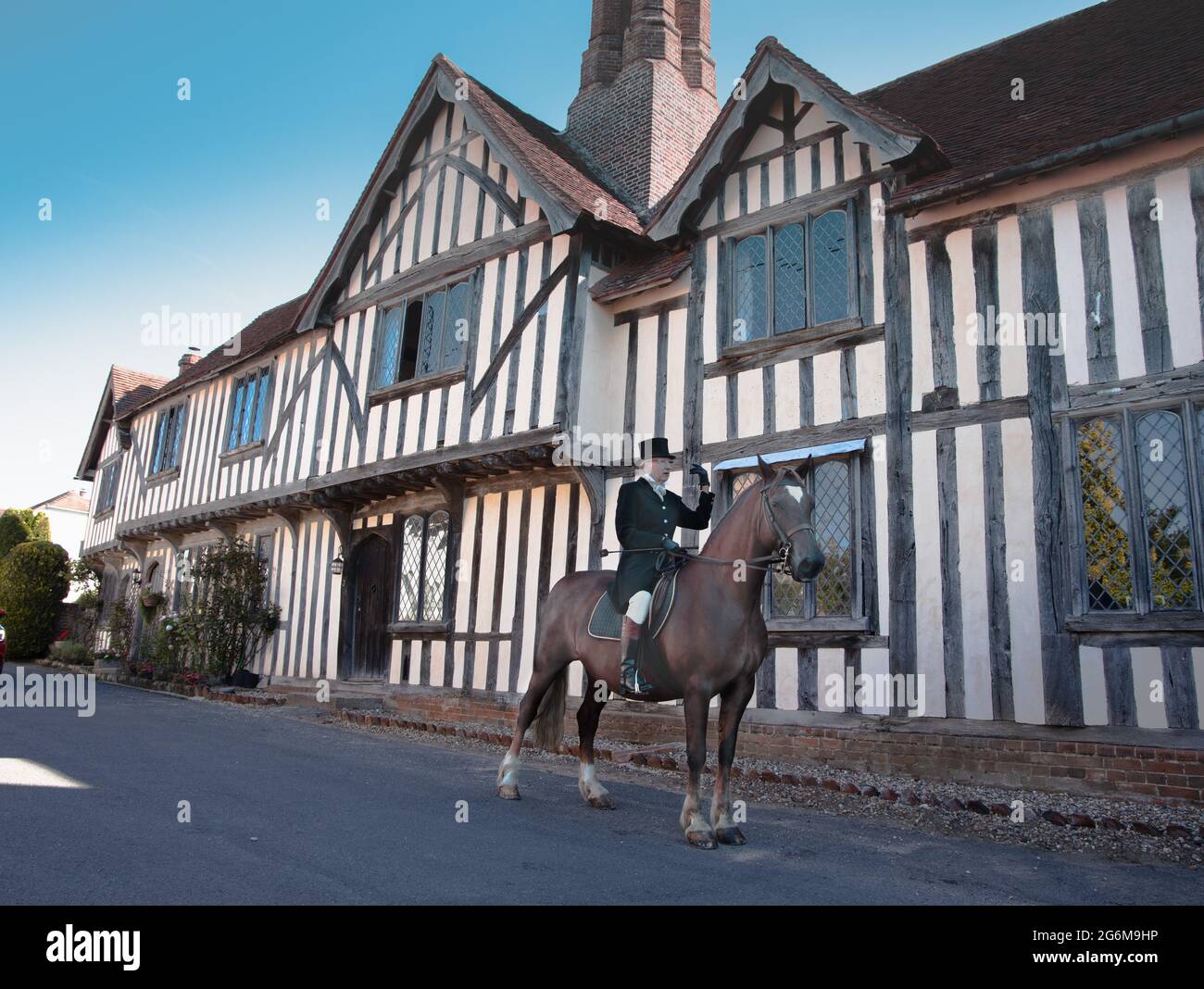 Lady formale di equitazione abitudine cavalcare un cavallo cacciatore di baia in un grazioso villaggio Suffolk di Nayland in Inghilterra con un vecchio edificio in legno sullo sfondo Foto Stock