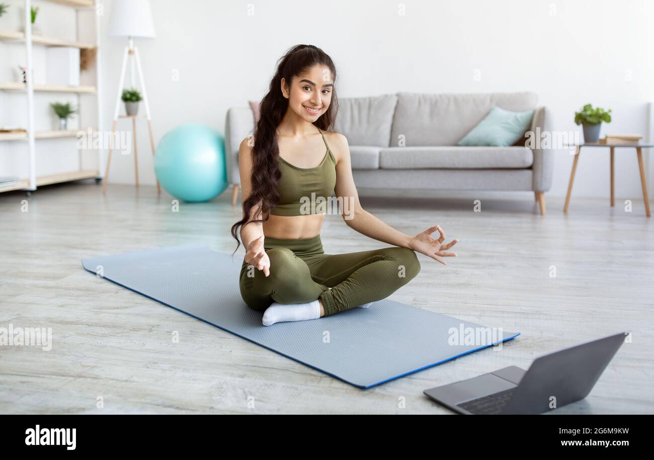 Una donna indiana pacifica che ha una meditazione online o una lezione di yoga, seduta in lotus posa di fronte al computer portatile a casa Foto Stock