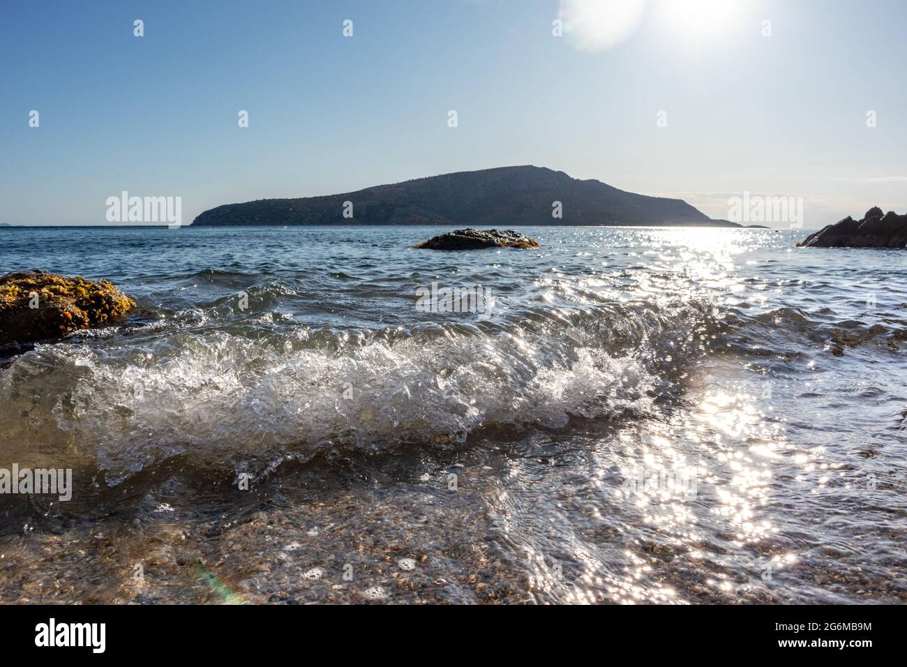 Il sole splende su onde bianche che colpiscono da vicino la costa selvaggia. Mar Mediterraneo con rocce, acqua limpida e fascio di sole. Viaggia in Grecia vicino ad Atene. Estate seasi Foto Stock