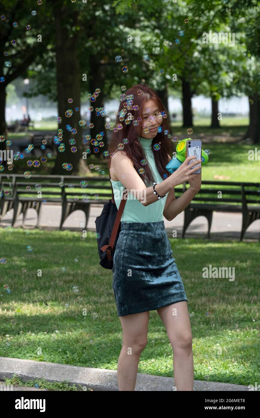 Una donna snella felice prende le immagini selfie che soffia le bolle da un thingie di plastica. In un parco a Queens, New York City. Foto Stock
