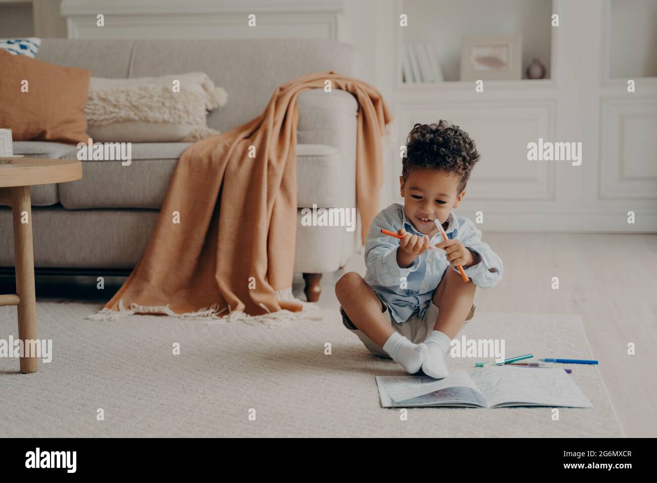 Carino capretto afro americano con libro da colorare seduto sul pavimento in soggiorno accogliente Foto Stock