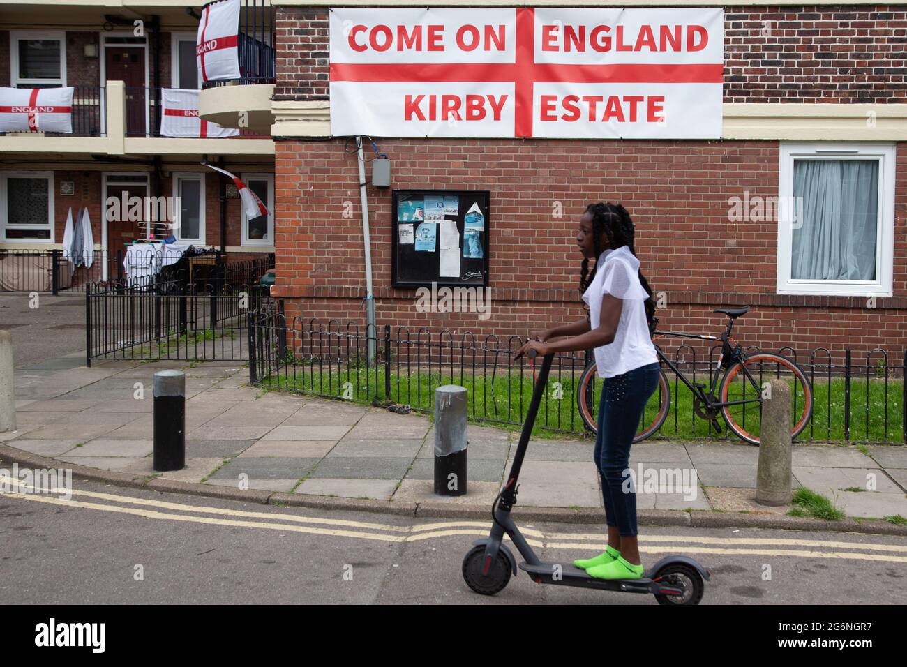 Una ragazza cavalcava uno scooter davanti alle bandiere inglesi e si accamparono drappeggiato sui balconi e attaccati alle pareti della tenuta Kirby a Bermondsey, a sud-est di Londra. Un'abitazione è stata coperta con bandiere inglesi prima della partita semifinale contro la Danimarca. Foto Stock