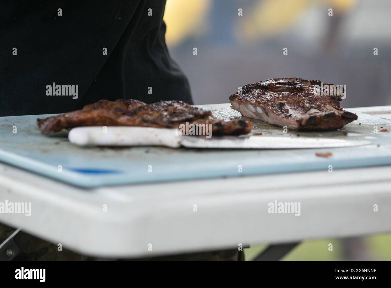 Un uomo che taglia le costole del barbecue dalla griglia fumatore sul tagliere con i guanti sopra e un coltello grande. Foto Stock