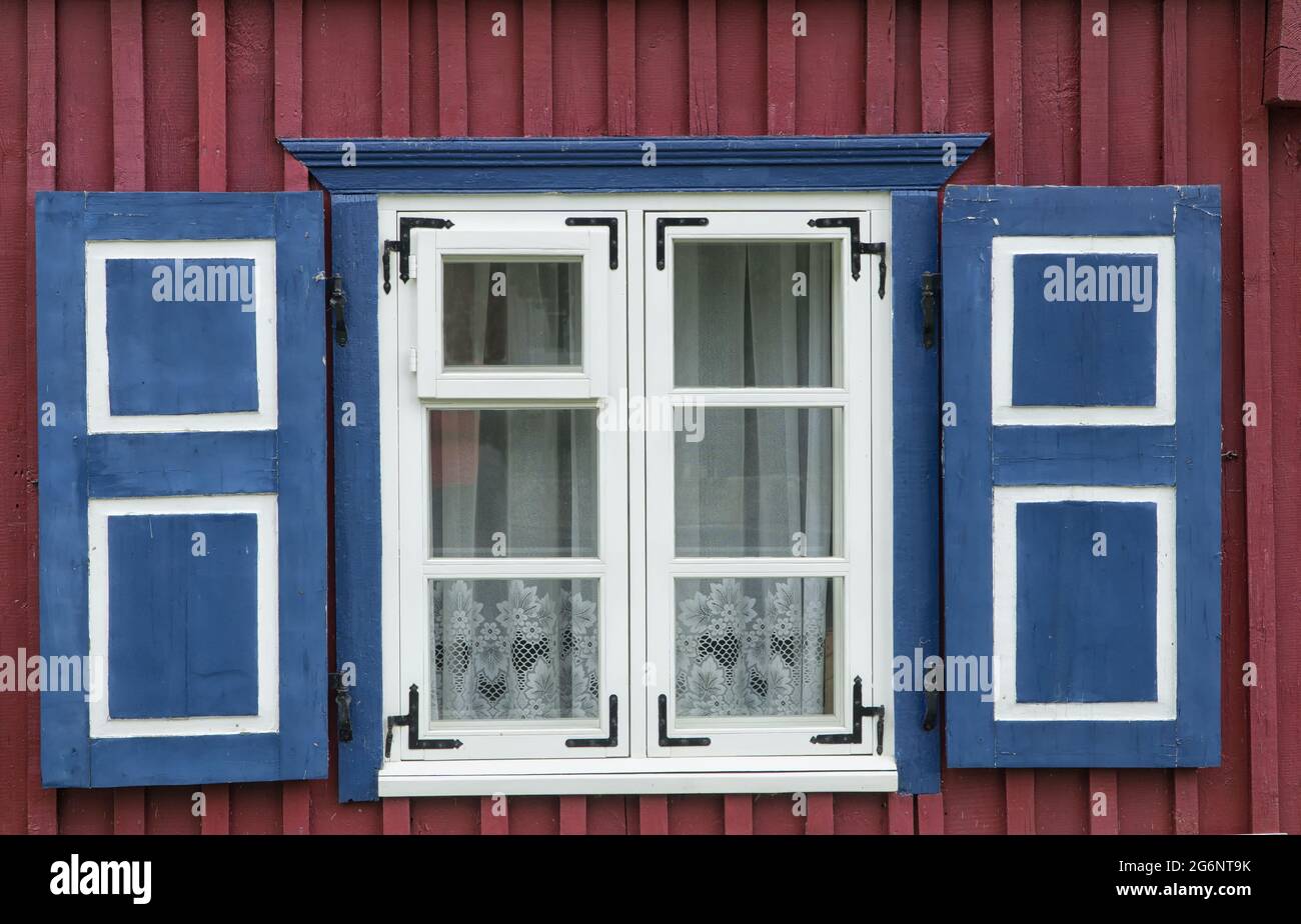 Finestra con cornice bianca e persiane blu sulla facciata rosso-marrone del tradizionale edificio in legno. Tenda in pizzo nella finestra. Vista esterna. Foto Stock