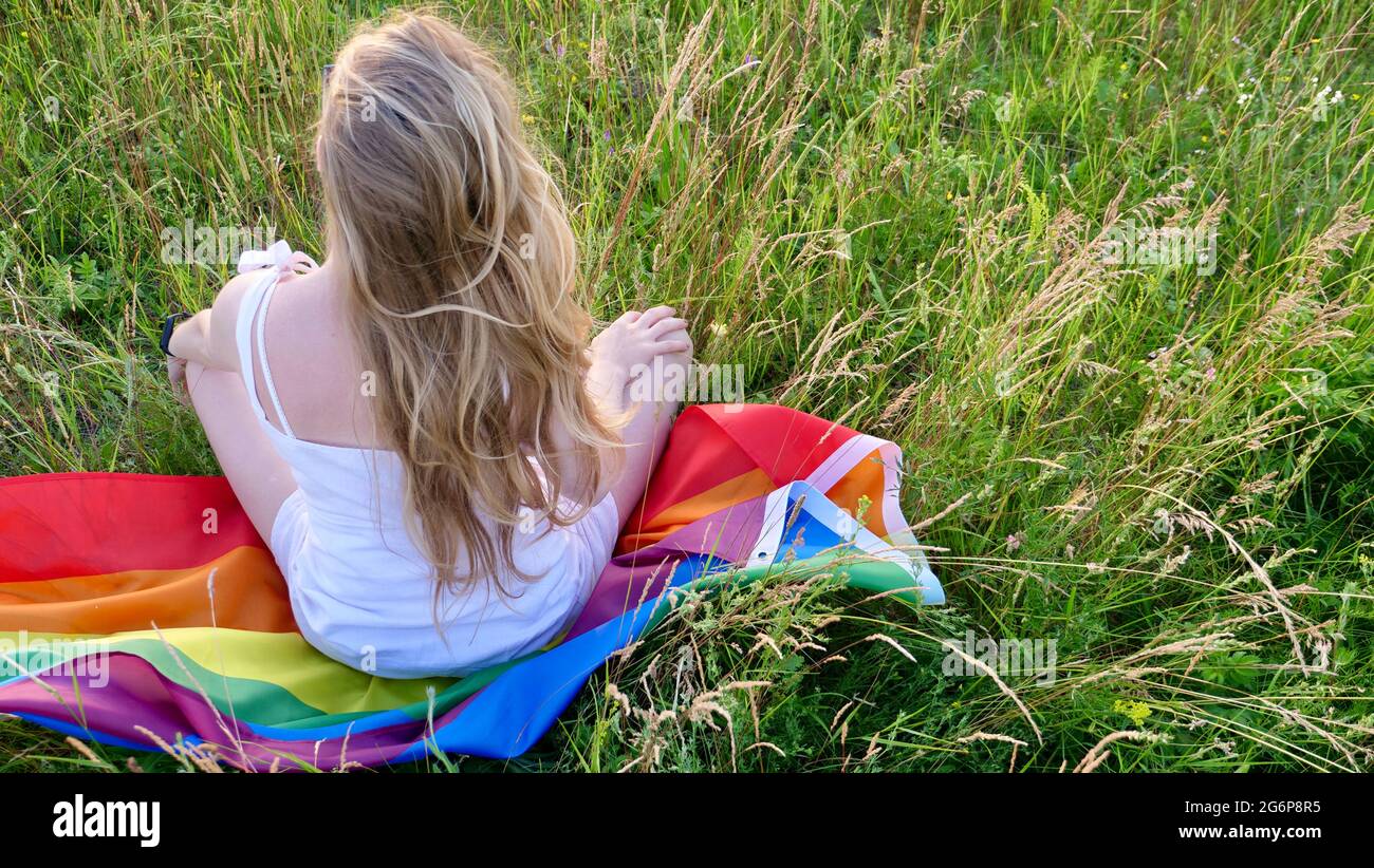 Donna bionda lesbica bisessuale seduta sulla bandiera LGBT sull'erba in abito bianco. Sostegno all'orientamento non tradizionale nel mese della dignità, giorno della bisessualità. Foto Stock