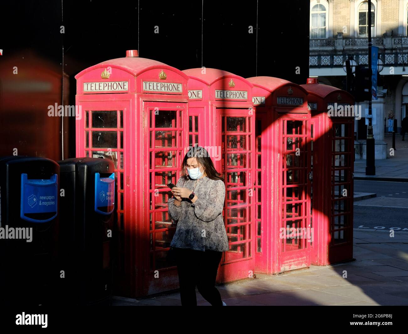 Una donna che indossa un facemask e guarda giù il suo cellulare passa una fila di scatole telefoniche nel centro di Londra in una soleggiata serata estiva. Foto Stock