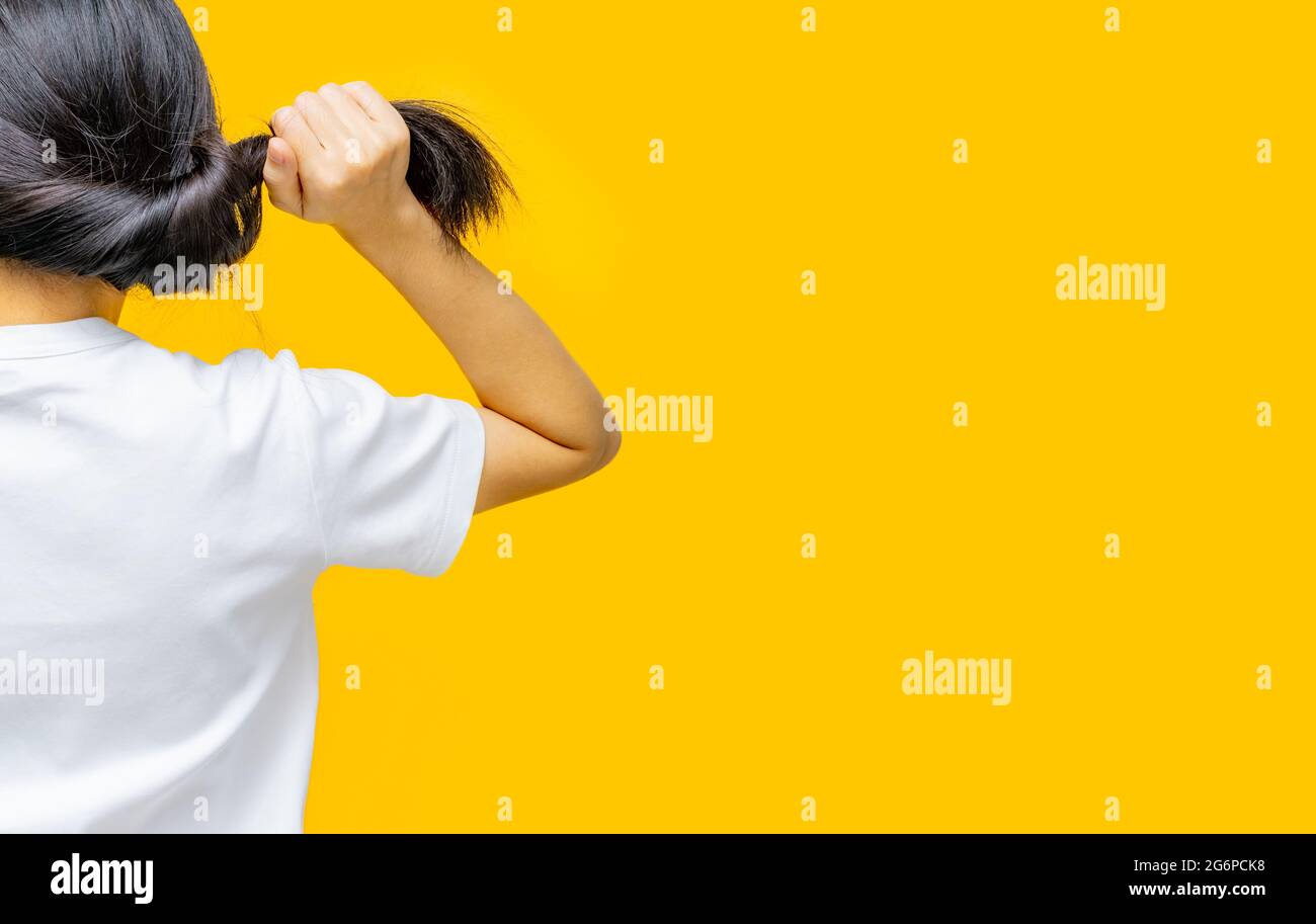 Vista posteriore della donna asiatica che tiene i capelli danneggiati su sfondo giallo. Perdita dei capelli e problema dei capelli sottili in donna. Capelli lunghi neri asciutti e fragili bisogno Foto Stock