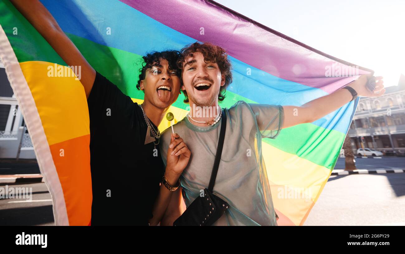 Giovani uomini non conformi che celebrano l'orgoglio gay. Due giovani gay sorridono allegro mentre alzano una bandiera di orgoglio arcobaleno. Due membri della com LGBTQ+ Foto Stock