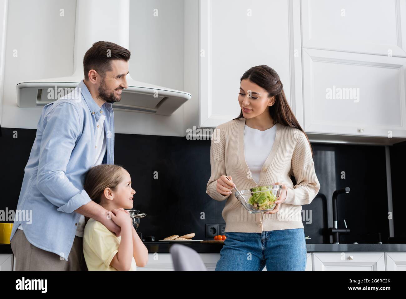 donna sorridente che tiene la ciotola di insalata di verdure vicino a marito e moglie in cucina Foto Stock