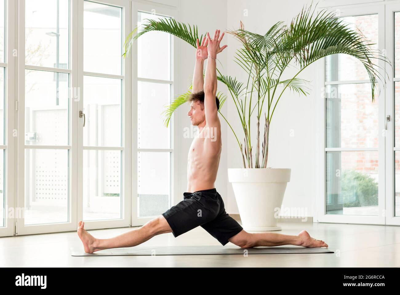 L'uomo che esegue un Hanumanasna o la posa di yoga delle scimmie che si divide in avanti con le braccia sollevate in una palestra di alta chiave con copyspace in un concep di salute e fitness Foto Stock