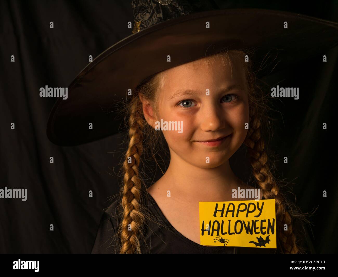 Piccola strega ragazza graziosa in cappello nero del mago tiene il saluto felice Halloween e ride. Sorridente bambino positivo vestito in costume da festa fantasma guardando Foto Stock