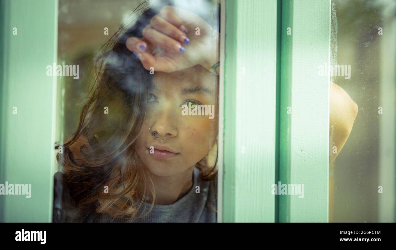 Una giovane donna, fotografata attraverso la finestra. Sta poggiando sul braccio sollevato contro la finestra. Foto Stock