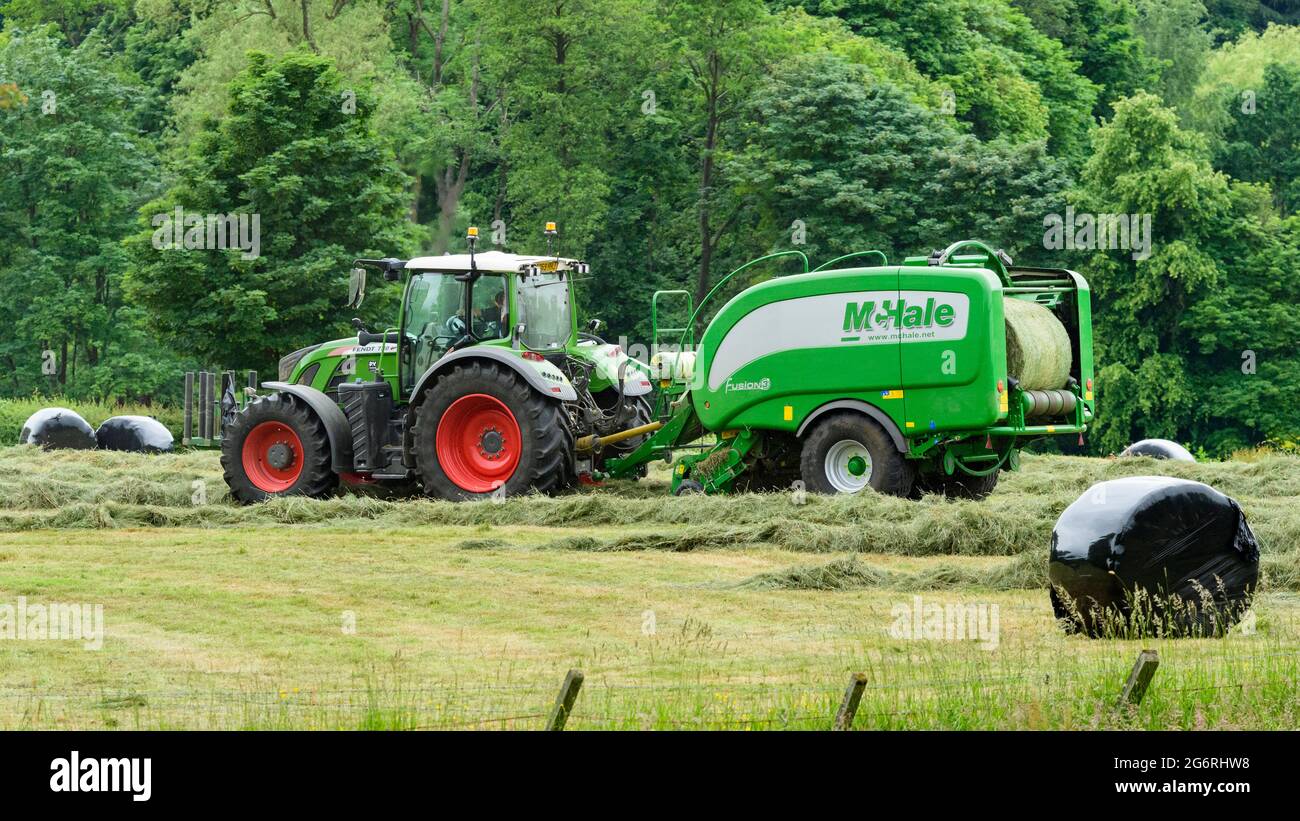 Produzione di fieno e insilato (coltivatore in trattore agricolo al lavoro in campo rurale, raccolta di erba secca tagliata, balla rotonda non imballata in imballatrice) - Yorkshire Inghilterra, Regno Unito. Foto Stock