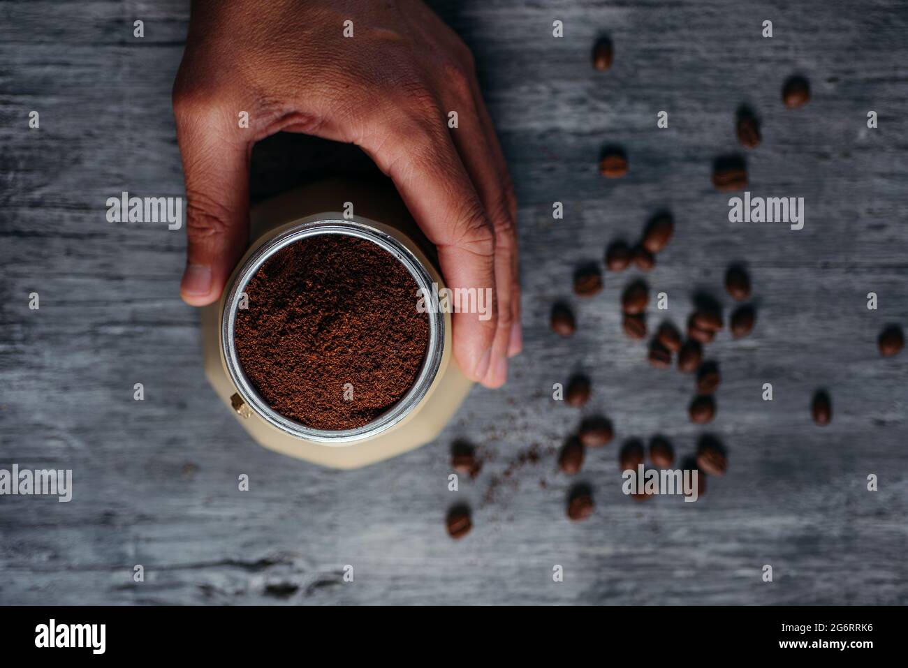 vista ad alto angolo di un giovane caucasico che sta preparando il caffè in una tazzina di moka beige, su un tavolo di legno rustico grigio cosparso di un po' di caffè arrosto Foto Stock
