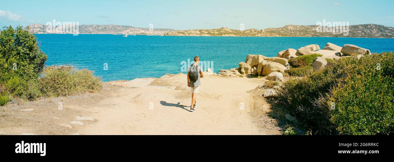 Un giovane, con uno zaino, cammina lungo la costa del Mediterraneo in Sardegna, Italia, con l'isola la Maddalena sullo sfondo, in una padella Foto Stock