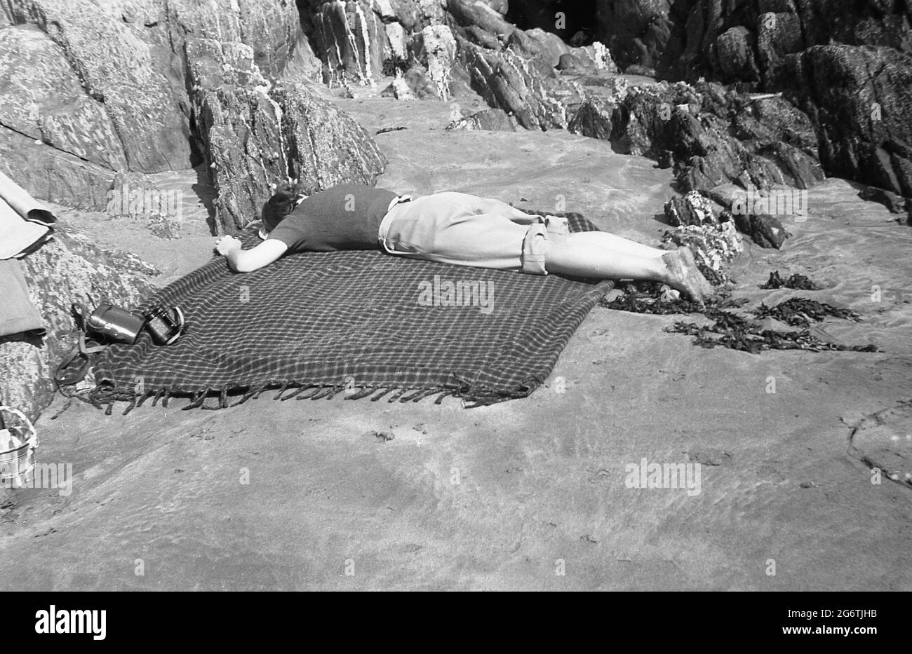 Anni '50, storico, una giovane donna con pantaloni arrotolati, sdraiata faccia in giù su un tappeto oltre ad alcune rocce su una piatta, piccola zona sabbiosa di una spiaggia che ha un riposo, Inghilterra, Regno Unito. Probabilmente esausto dopo aver costruito castelli di sabbia per i suoi figli. Foto Stock